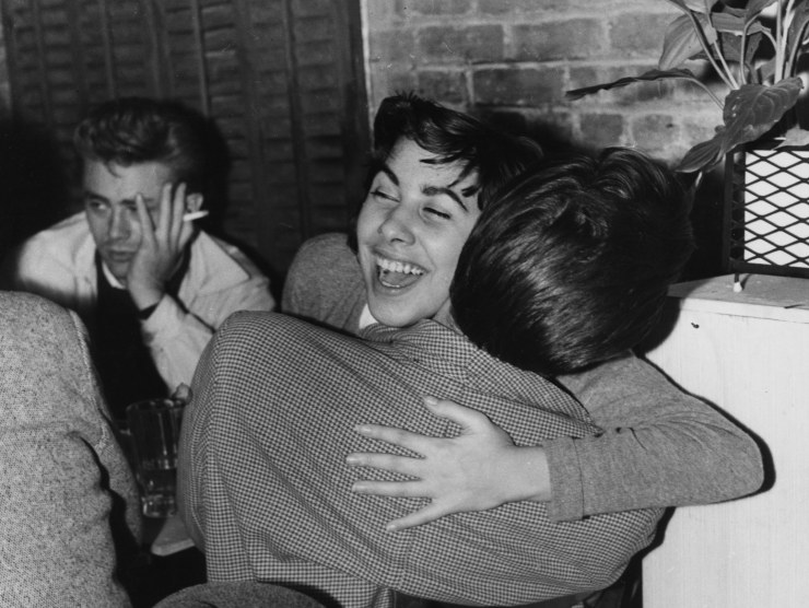 A young woman smiles as she hugs a man while actor James Dean smokes and looks away, bored, in this black and white photo.