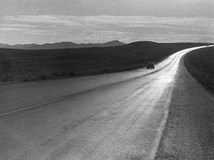A long curved roadway shimmers in the sun and a car drives towards the photographer with mountains in the background in this black and white photo.