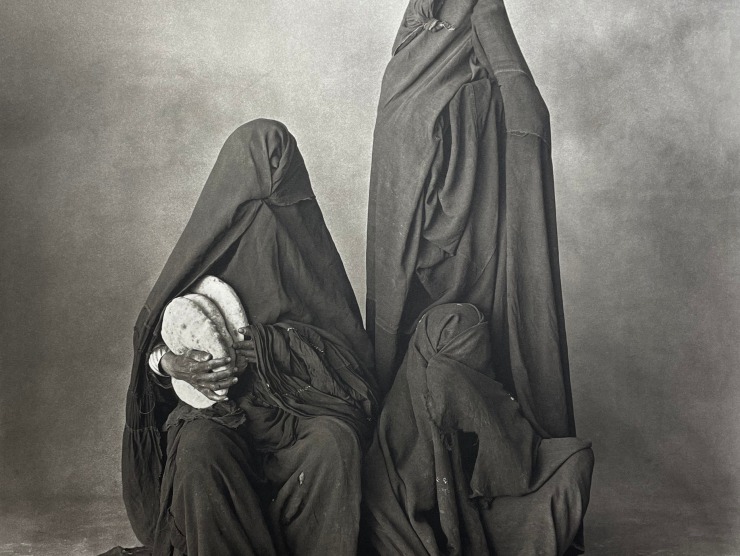 Three cloaked figures sit and stand against a grey backdrop while one person holds what appears to be bread in their hands in this black and white photograph.