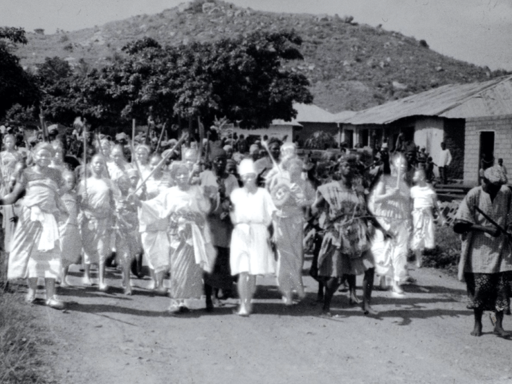 A procession of people walking down a dirt road is photographed in black and white.