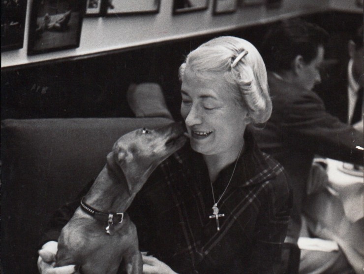 Black and white photograph of a woman at a restaurant table with a dog who is licking her face.