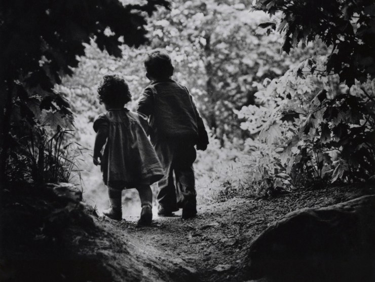 Two children are photographed from behind in black and white while they exit a forest and walk into a brighter landscape.