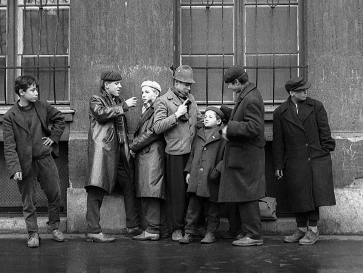 A group of boys wearing coats are photographed in black and white while they lean against a building wall outside.