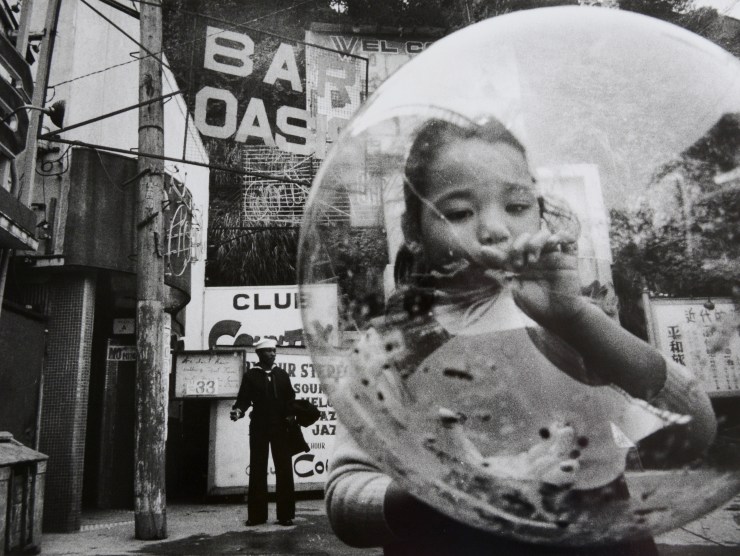 A child blows up a large, clear balloon and a sailor stands in the distance against a wall in a black and white photo