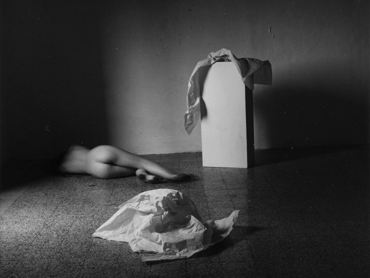 A young woman lays down while naked, facing away from the camera, and moves her feet, as wrinkled white tissue paper sits on the floor near her and on top of a white pedestal nearby in this black and white photo