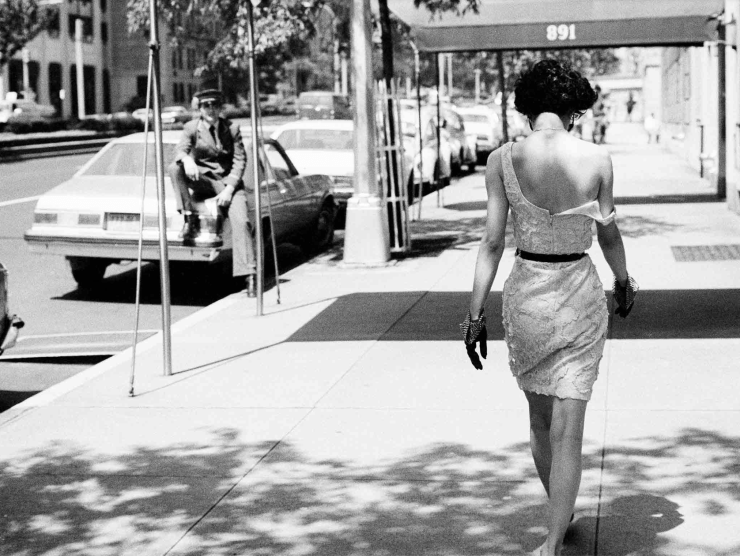 A woman is photographed from behind while wearing a short dress, one shoulder strap off her shoulder, and her head down, while a doorman sitting on the bag of a car looks at her in this black and white photograph.