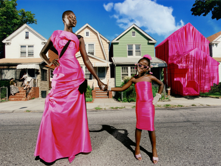 A tall Black female model stands in a hot pink formal dress on a street with houses, while holding the hand of a little girl's hand. The girl is also wearing hot pink and one of the houses in the background is draped in hot pink fabric.