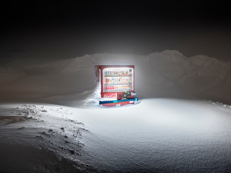 A vending machine sits in deep snow and is photographed while glowing at night.