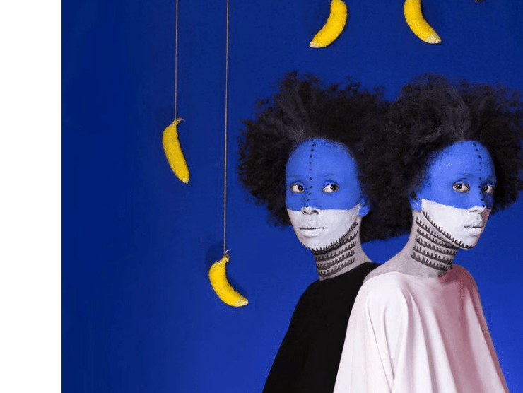 Two women standing close to each other have blue and white face paint and stand in front of a blue wall with bananas dangling down from above.