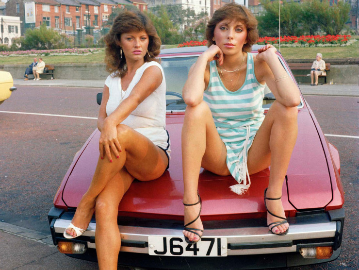 Two stylish women sit on the hood of a red car.