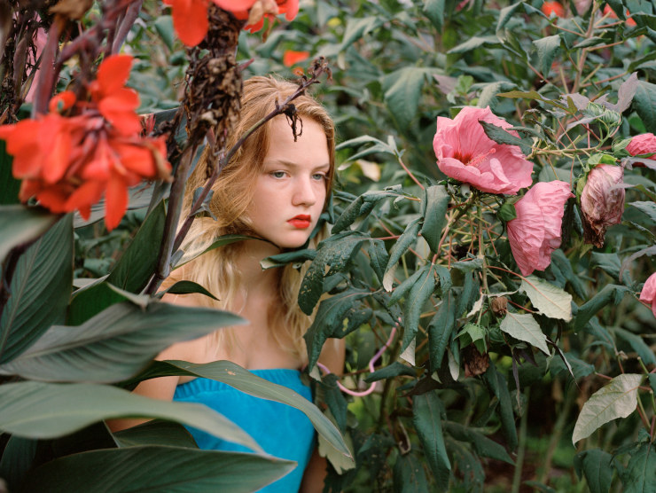 A young girl in a teal romper wears bring reddish-orange lipstick and sits amongst tall colorful flowers while staring into the near-distance.