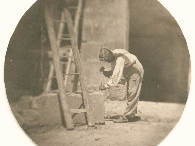 A man bends to use a hammer and chisel on a large stone block in this vintage black and white photograph