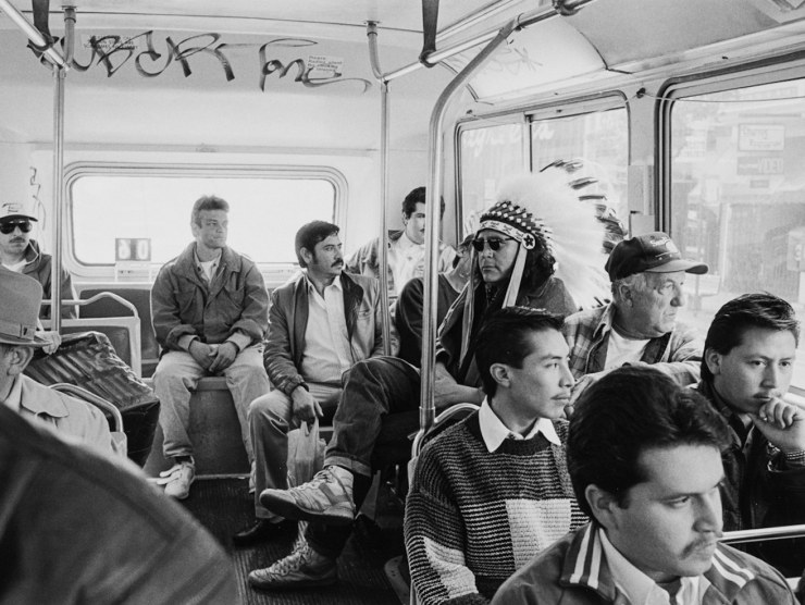 An indigenous man in a Native American feathered headress sits on a crowded bus while wearing sunglasses in this black and white photo.