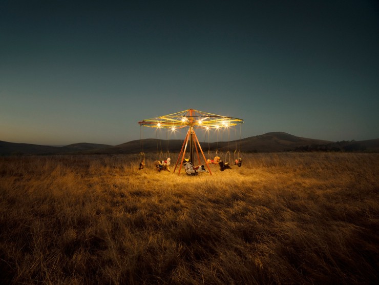 A suspension carousel is lit from above and situated in a wild field in front of mountains, photographed at dusk in color.