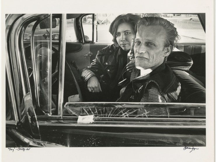 A couple sit in an old car while wearing leather jackets and looking towards the photographer in this black and white photograph.