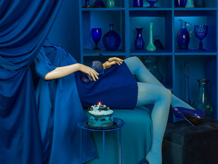 A partially obscured woman is shown wearing all blue next to a birthday cake with lit candle in an otherwise blue scene.
