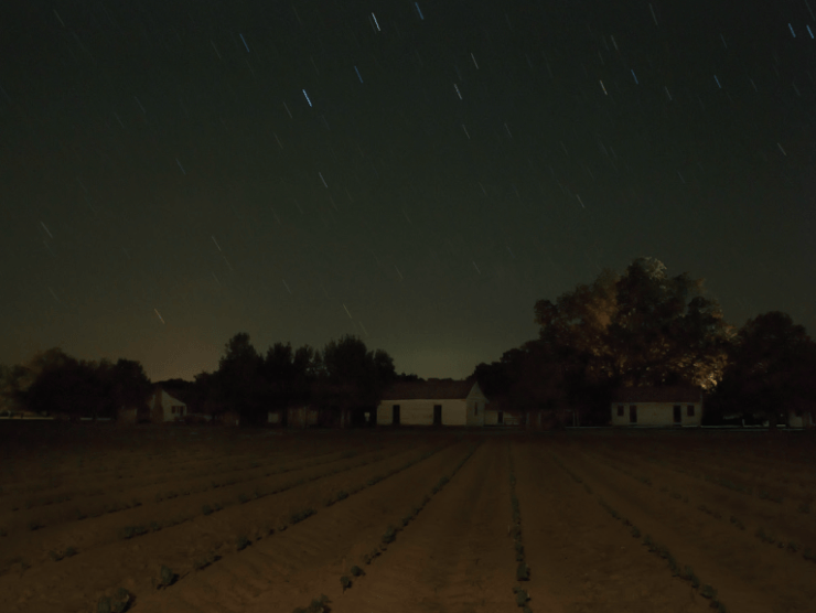 Photographic artwork that shows a farm field at night with houses and a dark star-lit sky above