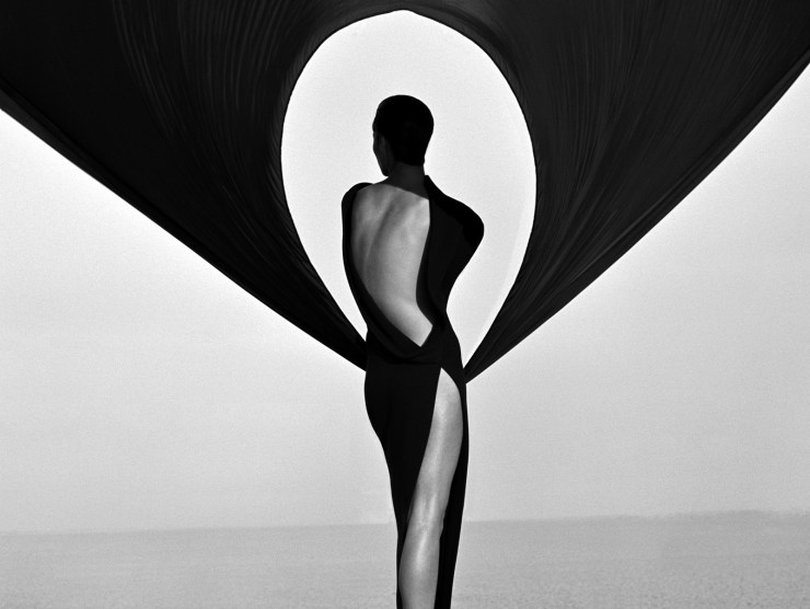 A black and white photo of a figure in a black dress in the desert with black fabric suspended overhead