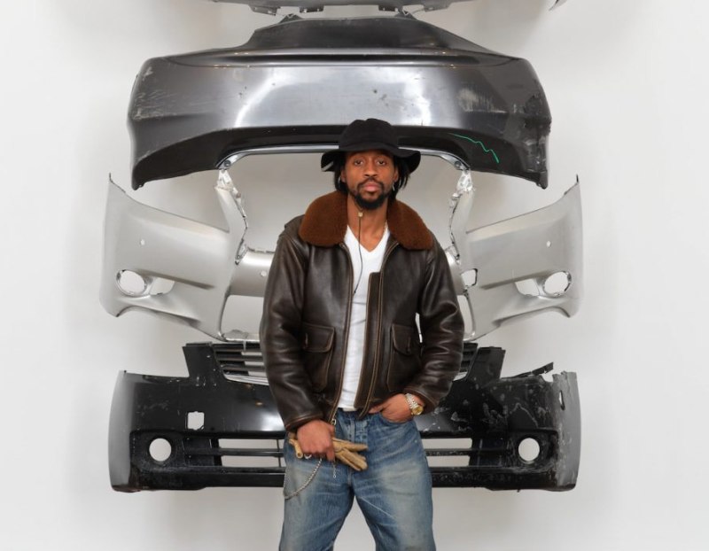 Artist Daniel Tyree Gaitor-Lomack, a Black man wearing a hat and brown leather jacket, stands in front of an installation comprised of the front bumpers of cars displayed in a column on a wall.