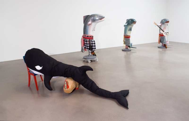 Installation image of a killer whale soft sculptures with three plastic mackrel sculptures in the background. Each mackrel is robed in gingham fabrics, one has a ukulele and another has a white electric guitar.