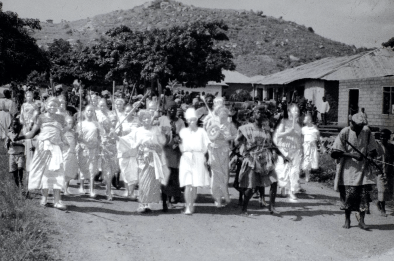A procession of people walking down a dirt road is photographed in black and white.