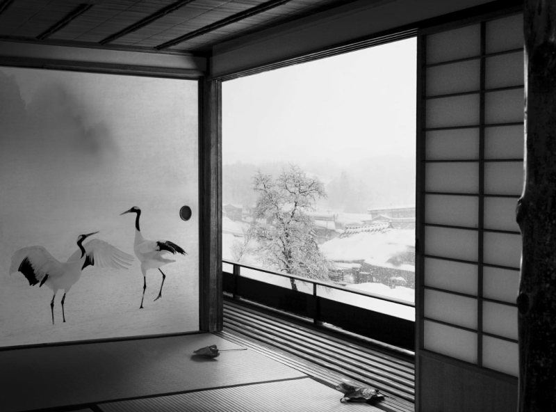 A black and white interior image shows a shimmering painting of cranes and a window with a snow scene outside.