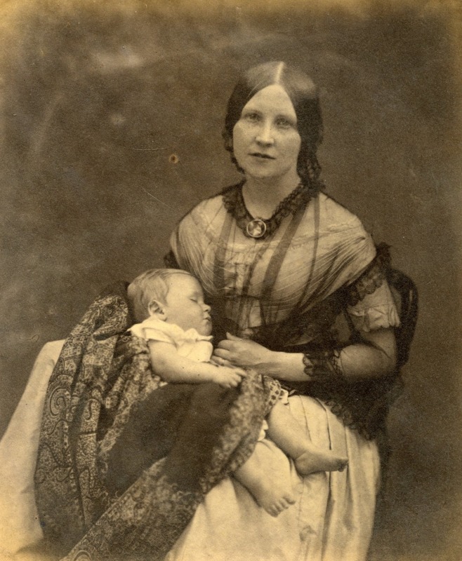 A woman wearing formal clothes poses in this sepia photograph while holding a baby.