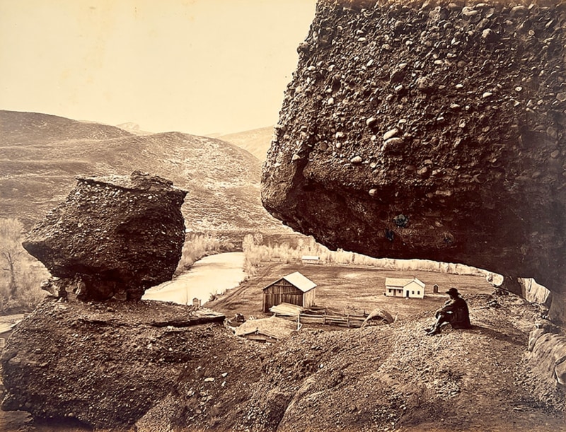 A man sits under a large boulder on a hilltop overlooking a barn and a house in this sepia photograph.