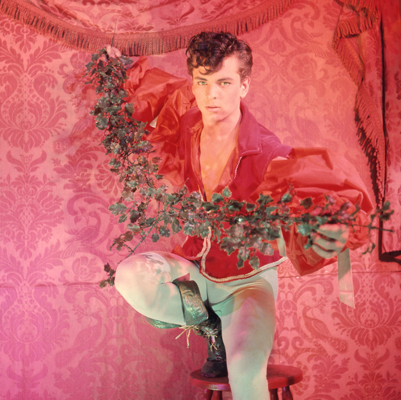 A man wearing red and green holds mistletoe in front of an ornate red wall