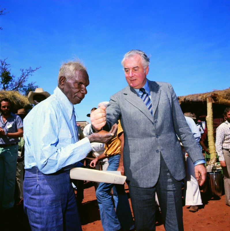 An older white man pours earth into the palm of an older black man.