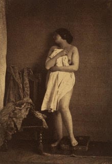 A nude woman stands holding a white cloth over her body while looking away from the camera in this sepia-toned print.