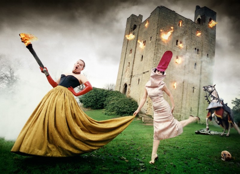 Two formally dressed people stand on the grounds of a burning castle with a white horse rearing behind them, and one figure holds a flaming torch