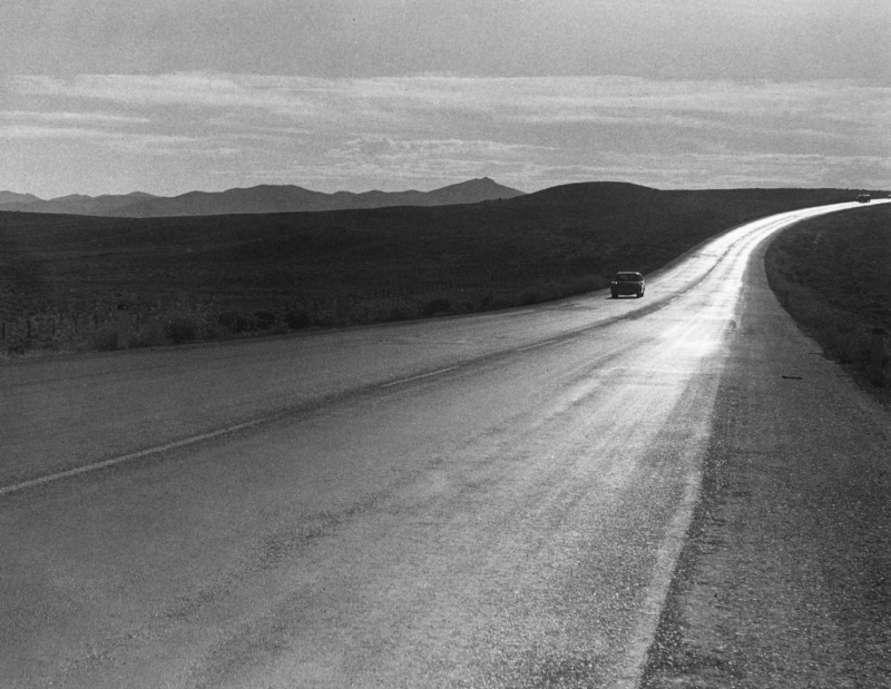 A long curved roadway shimmers in the sun and a car drives towards the photographer with mountains in the background in this black and white photo.