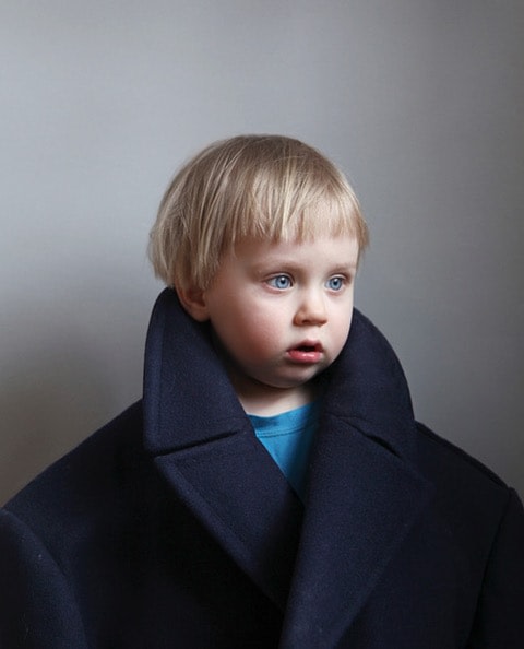 A young blond child wears an oversized coat in this color portrait.