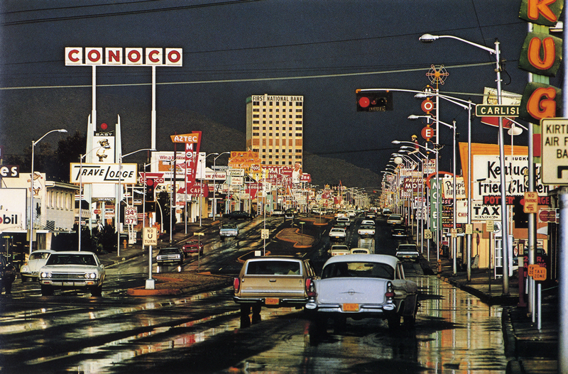 A colorful vintage photograph showing a roadway with lots of colorful signs on both sides of the road.