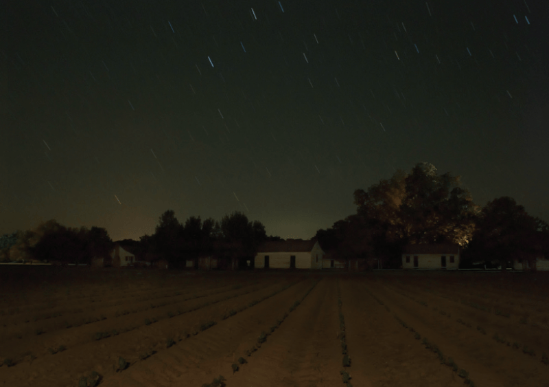 Photographic artwork that shows a farm field at night with houses and a dark star-lit sky above