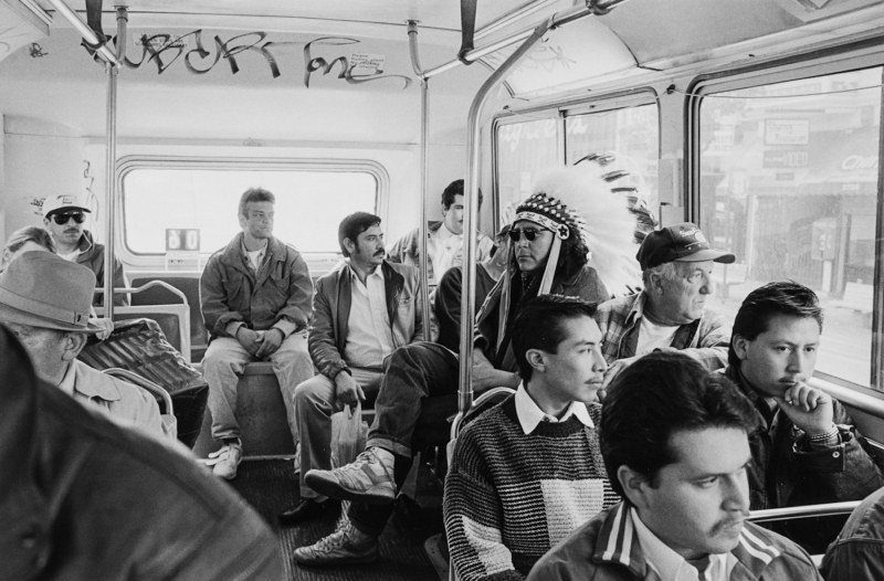 An indigenous man in a Native American feathered headress sits on a crowded bus while wearing sunglasses in this black and white photo.