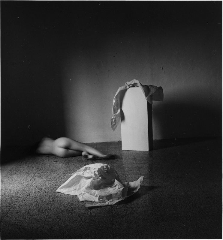 A young woman lays down while naked, facing away from the camera, and moves her feet, as wrinkled white tissue paper sits on the floor near her and on top of a white pedestal nearby in this black and white photo