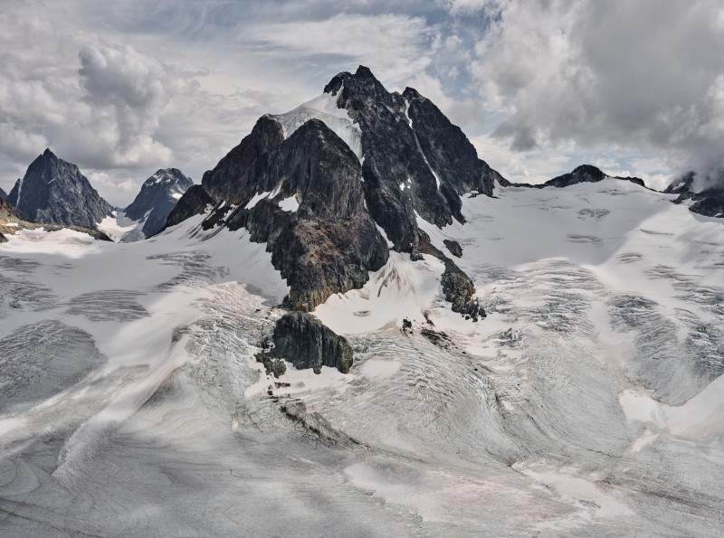 A mountain is photographed in sharp detail with snow melting off its peak and clouds surrounding in a pale blue sky.