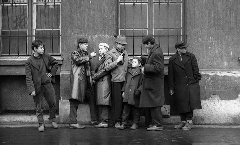 A group of boys wearing coats are photographed in black and white while they lean against a building wall outside.