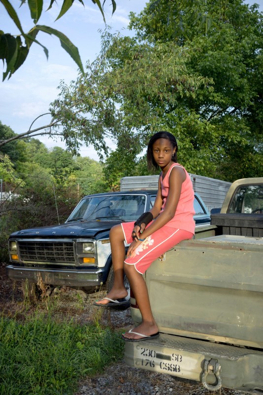 A color photo with a Black child wearing pink while sitting on the back of a truck and looking into the camera