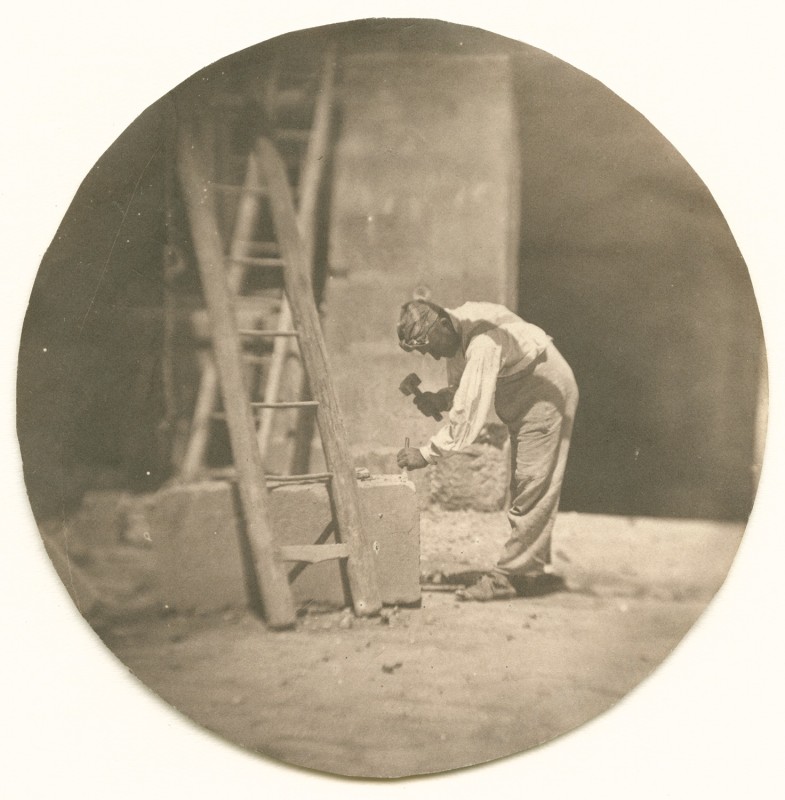 A man bends to use a hammer and chisel on a large stone block in this vintage black and white photograph