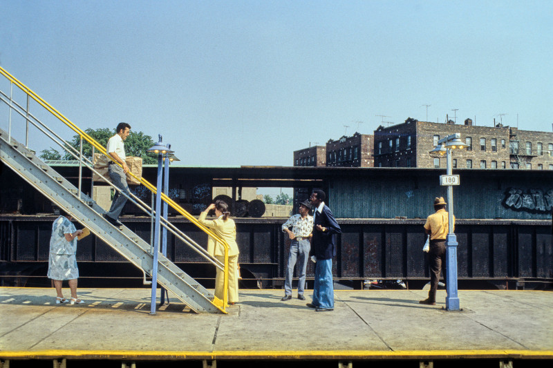 Several people wearing blue and yellow wait for the above-ground Subway in a scene with a blue sky and a yellow painted line in it.