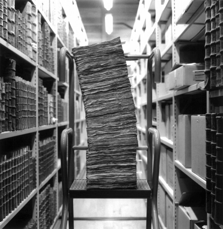 A stack of leaning papers in a library aisle, photographed in black and white