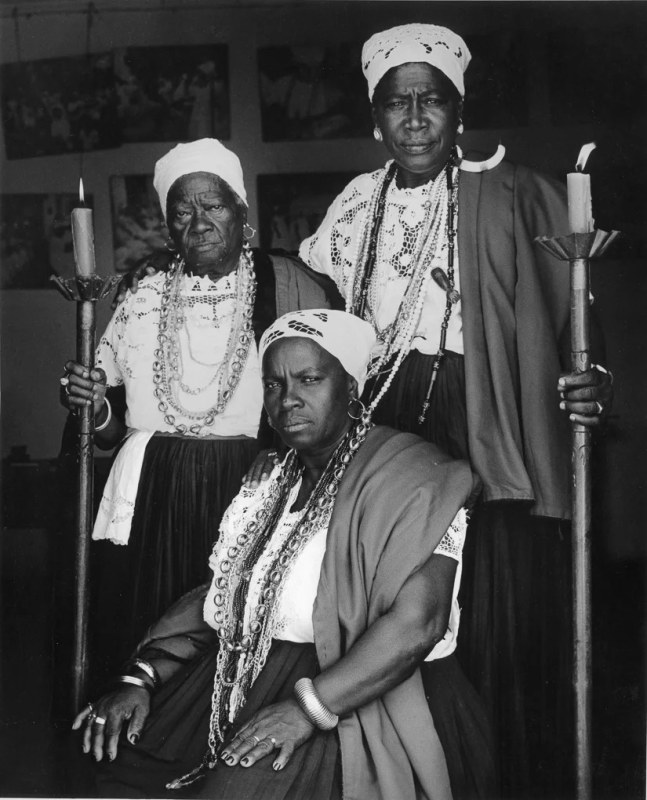 Three Black elders wear ceremonial dress and two hold candles