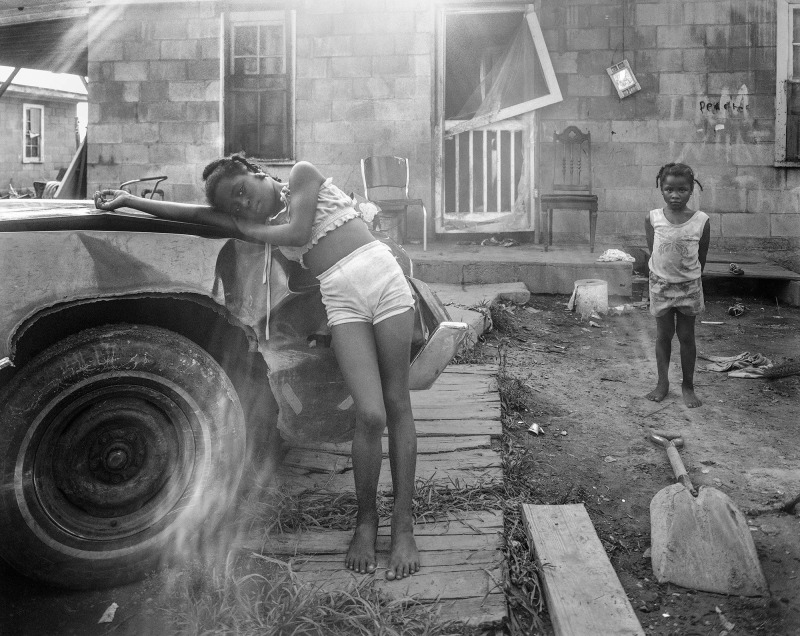 A Black child leans against a wrecked car in a yard while another child also looks at the camera