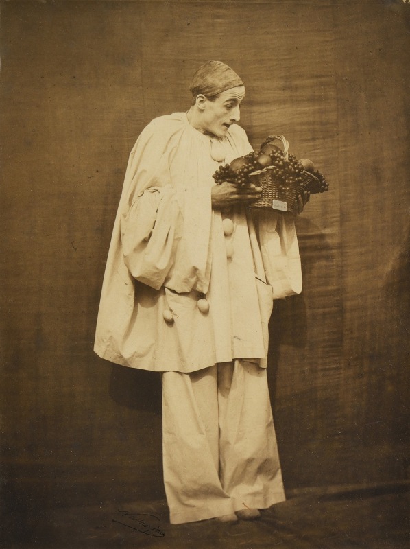 Pierrot the clown holds a basket of fruit while looking down at it in this sepia photograph.