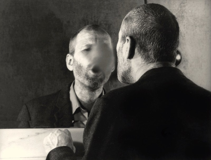 An unshaven man blows air onto a mirror, partially obscuring his face, in this black and white photograph