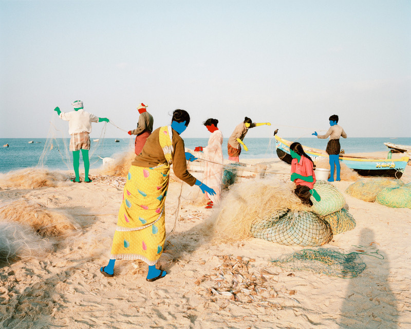 A group of people fish by the sea shore, holding nets, but their skin has been changed to bright primary colors such as blue, green, and red.