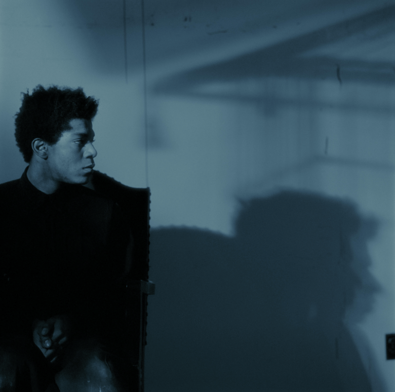 A cool-toned portrait of Jean-Michel Basquiat in profile with his shadow also showing in the corner of the image.